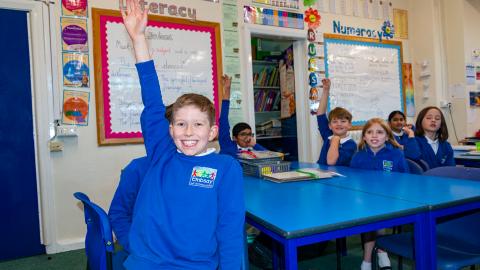 Pupils in a classroom setting, boy in the foreground confidently holds up his hand to answer a question